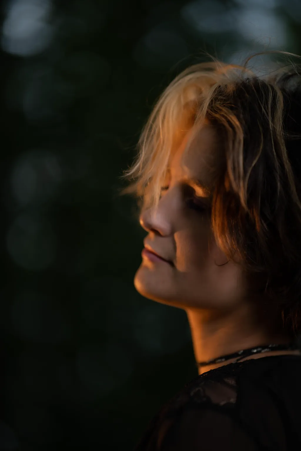 Side profile of a woman with eyes closed in golden light against dark woodland, Luxembourg