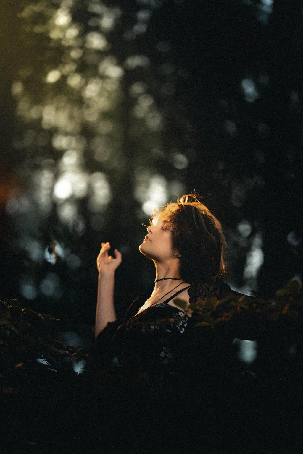 Woman in a floral black dress lifting her face to the light among woodland greenery in Luxembourg