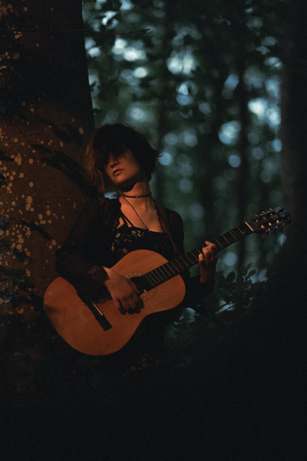 Woman playing an acoustic guitar against a tree trunk in dim Luxembourg forest light