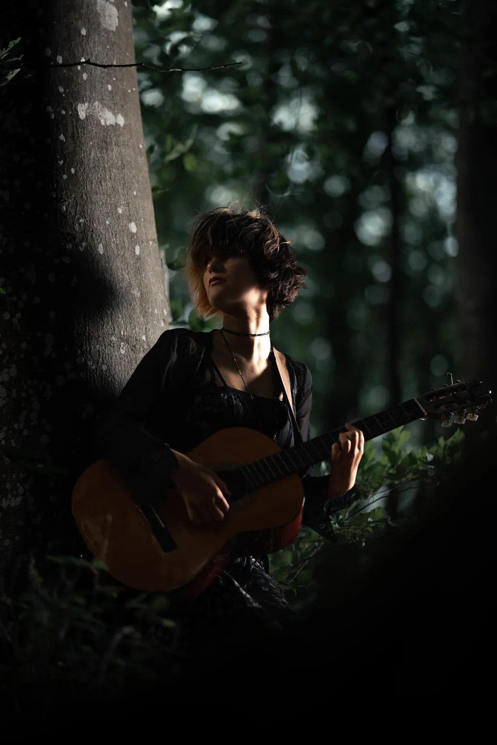 Guitarist leaning on a tree, lit by dappled woodland light in a Luxembourg outdoor portrait