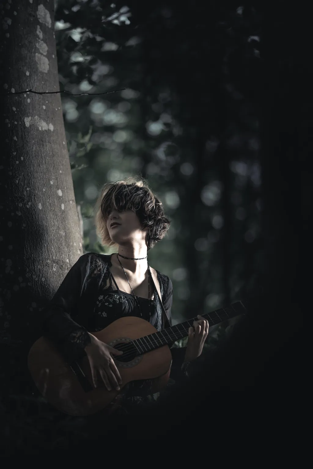Woman with guitar resting against a tree, face raised to forest light in Luxembourg