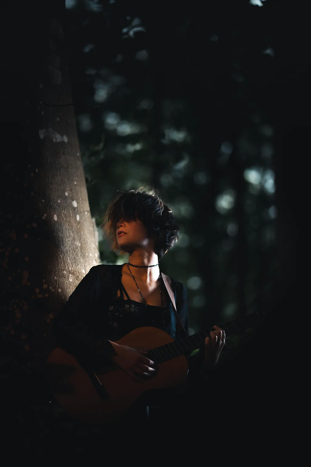 Woman playing guitar lit by a narrow beam of light in dark Luxembourg woodland