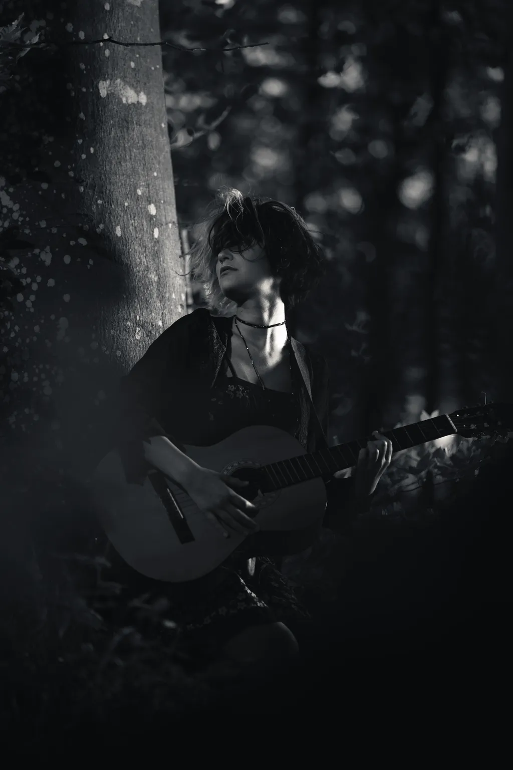 Moody monochrome portrait of a woman playing guitar beside a tree in a Luxembourg forest