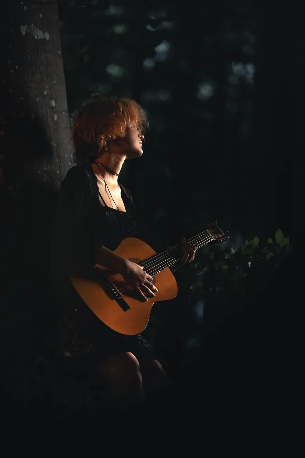 Seated woman playing acoustic guitar against a tree in deep shadow, Luxembourg woodland portrait