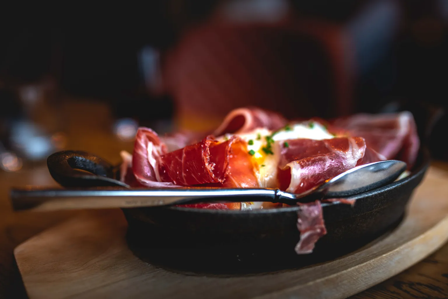 Baked egg dish draped with Iberian ham in a cast-iron pan, served on wood with a spoon