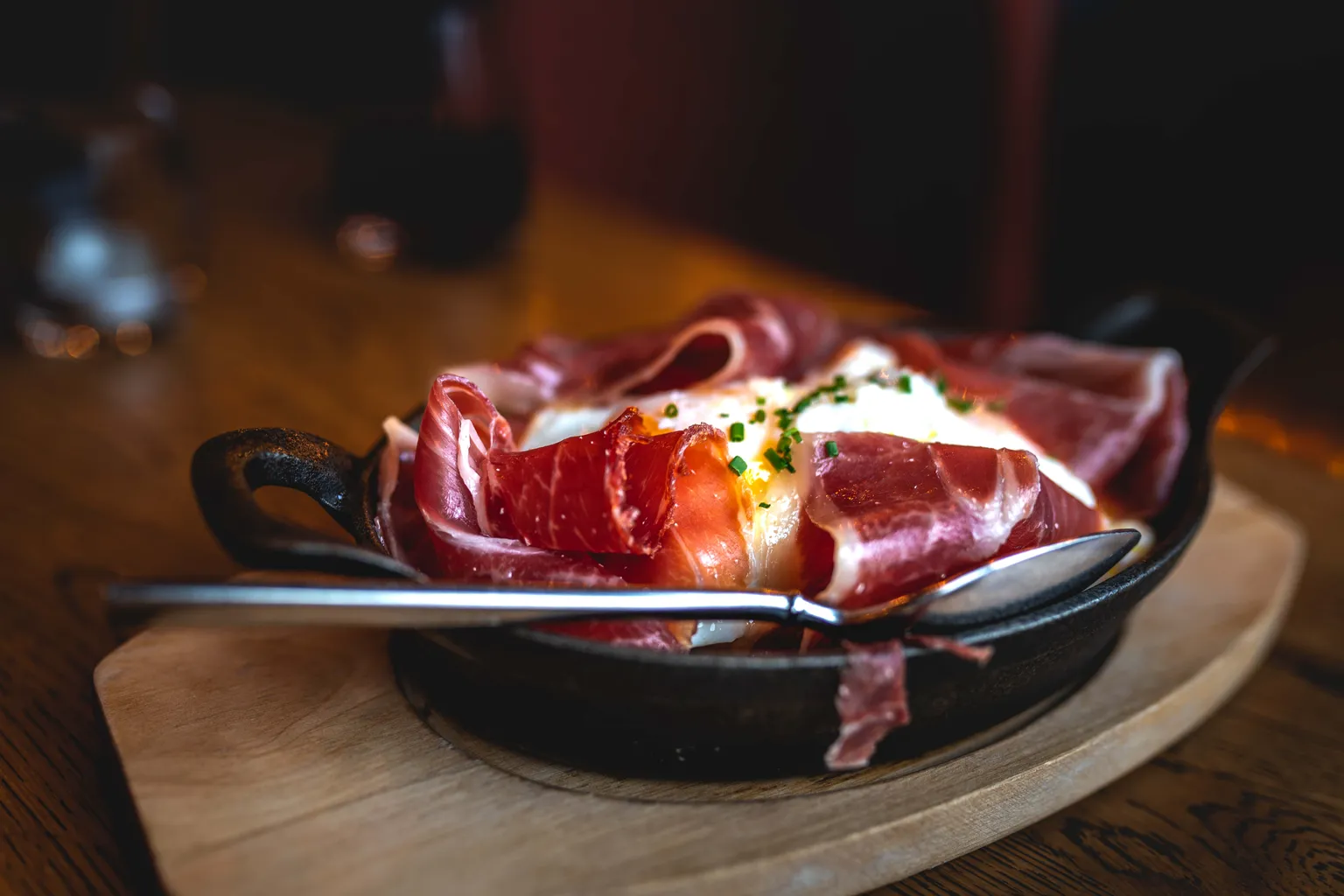 Cast-iron skillet of baked egg with cured ham and chives on a wooden board, warm restaurant ambience