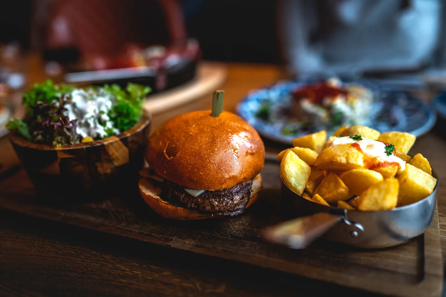 Restaurant table spread: burger, leafy salad bowl and copper pot of fried potatoes on a wooden board in Luxembourg