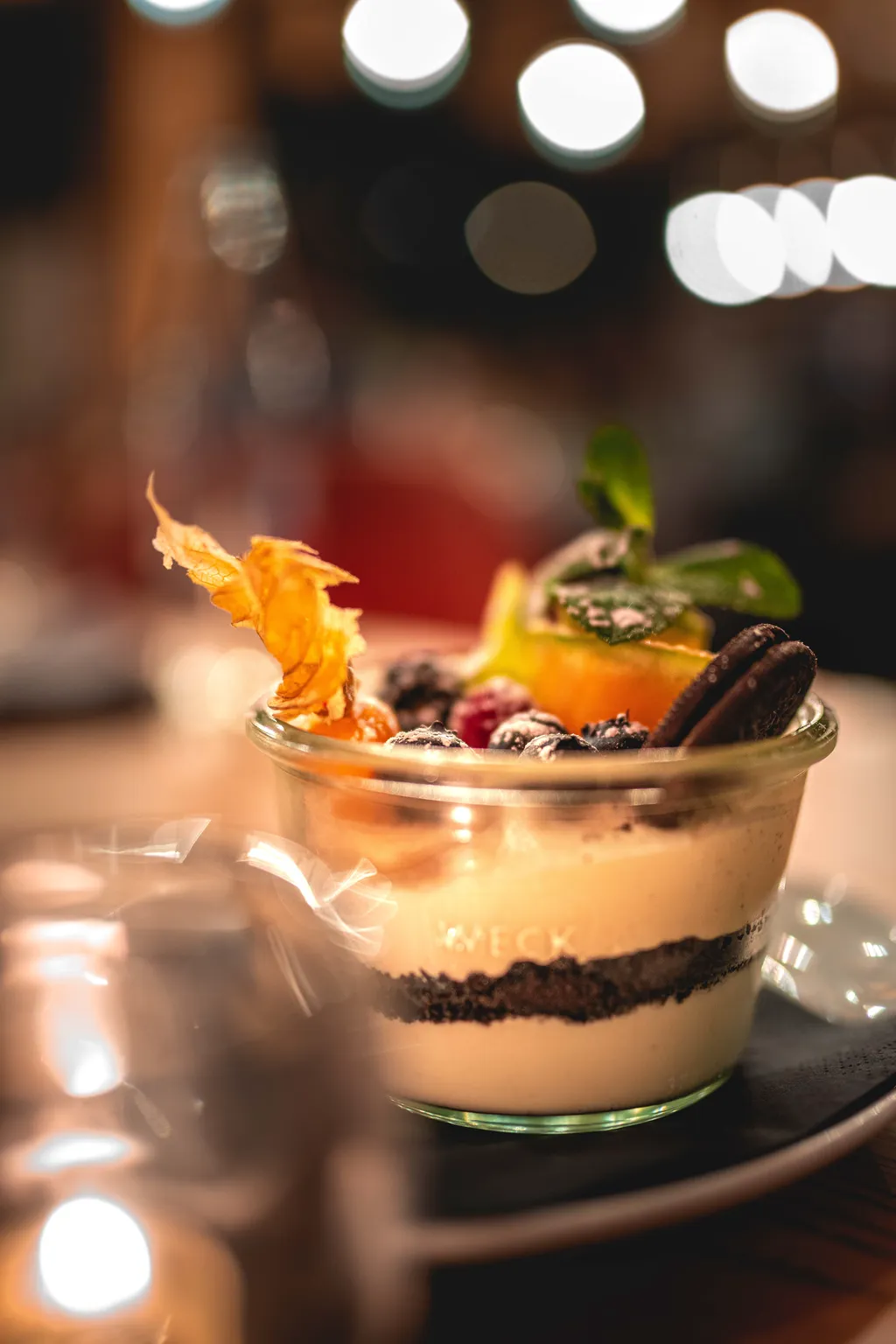 Weck-jar dessert with cookie-crumb layers, physalis and fresh berries against warm restaurant lights