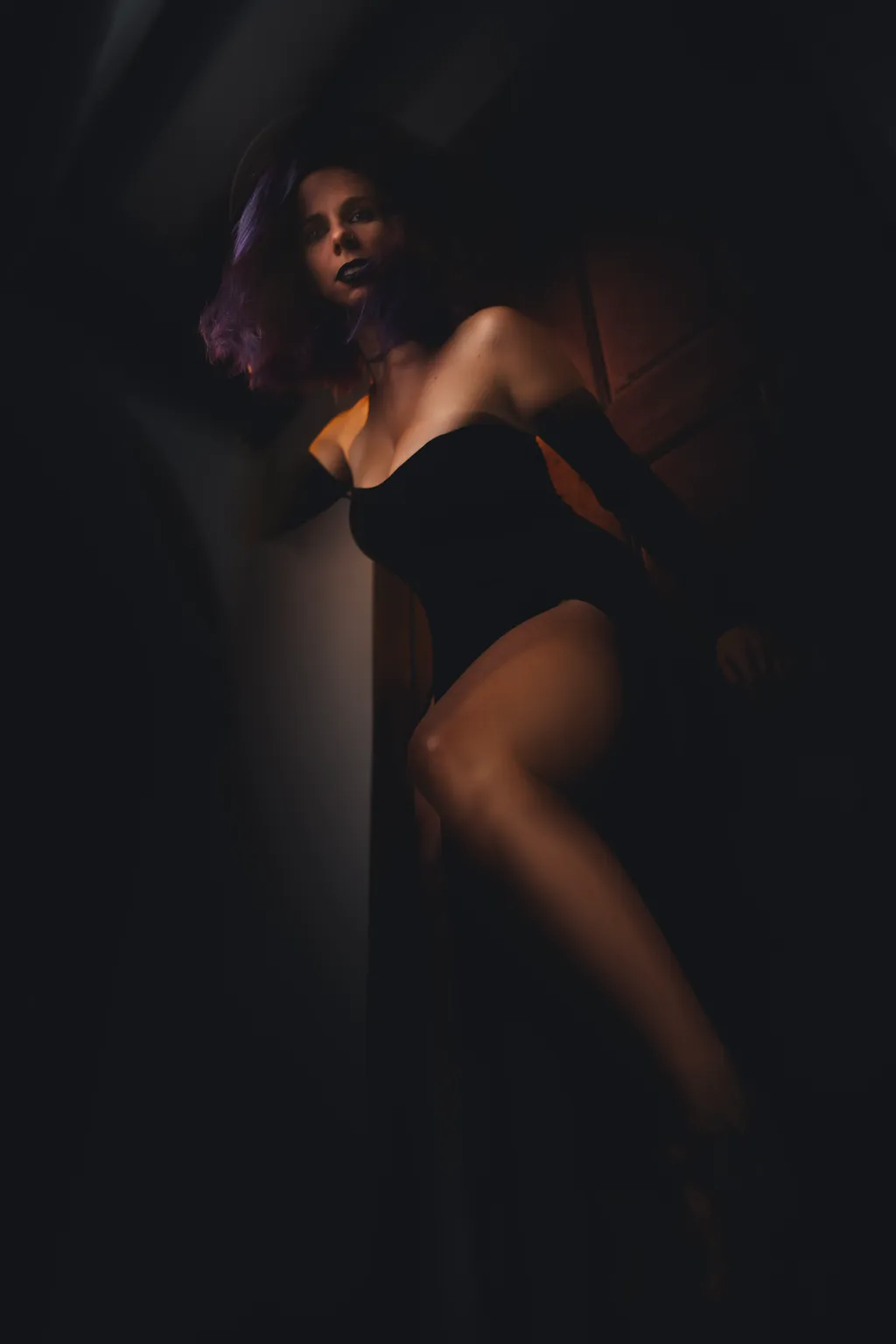 Low-key editorial portrait of a woman in a black bodysuit, warm amber light grazing her hair against near-total darkness.