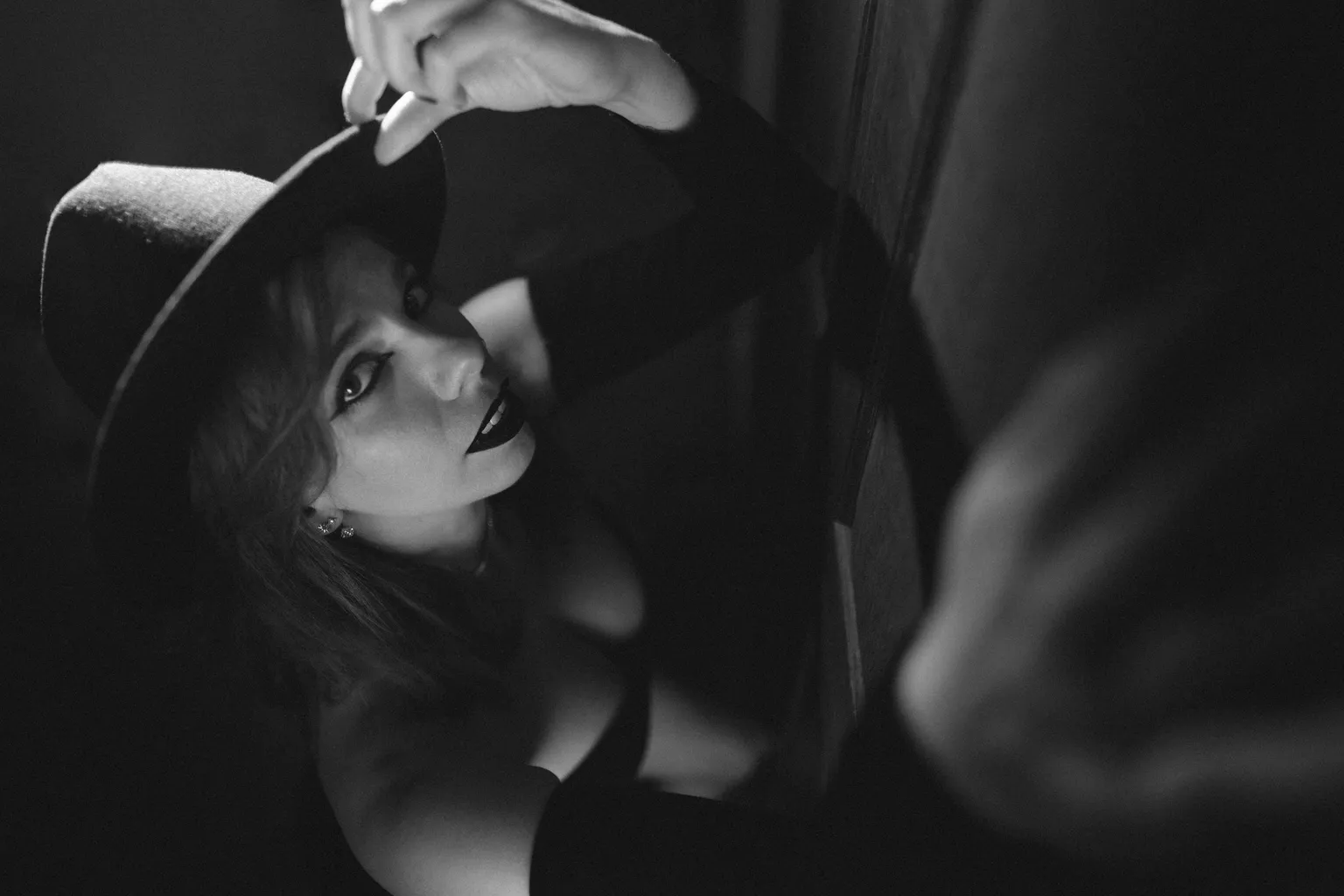 High-contrast noir portrait: a woman in a wide-brim hat tilting it back, red lips catching the light in deep shadow.
