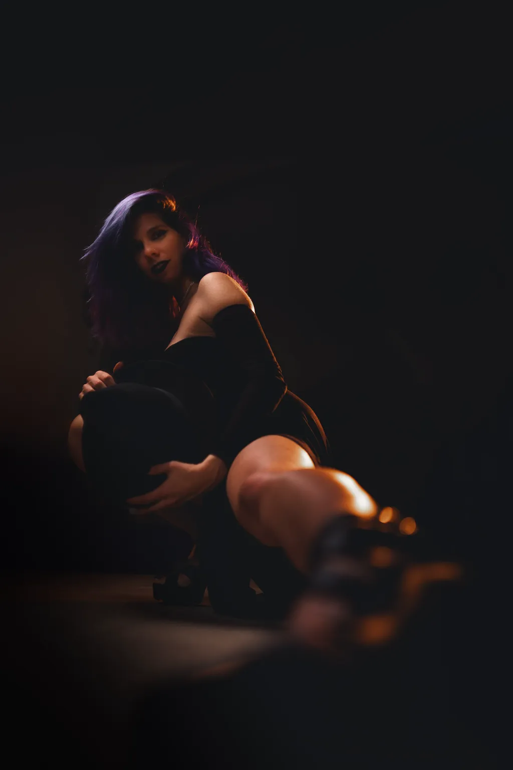 Cinematic low-key portrait of a seated woman in a black dress, warm light pooling on her skin and heeled shoe.