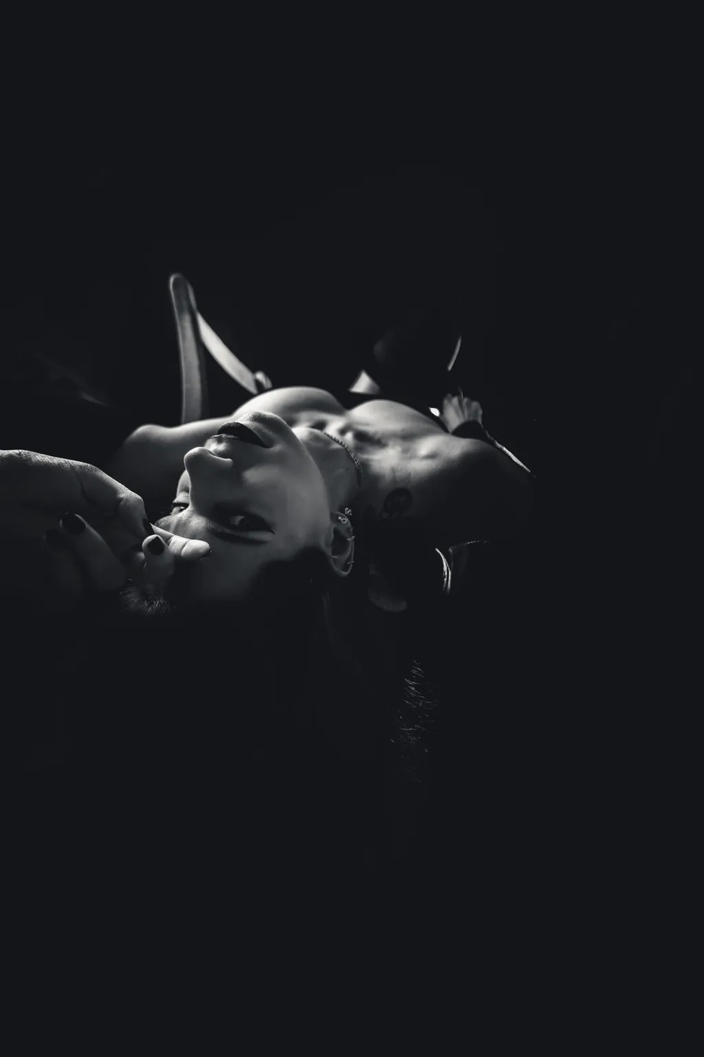 Sensual noir editorial portrait: a woman lying back with a hand near her face, lit dramatically against shadow.