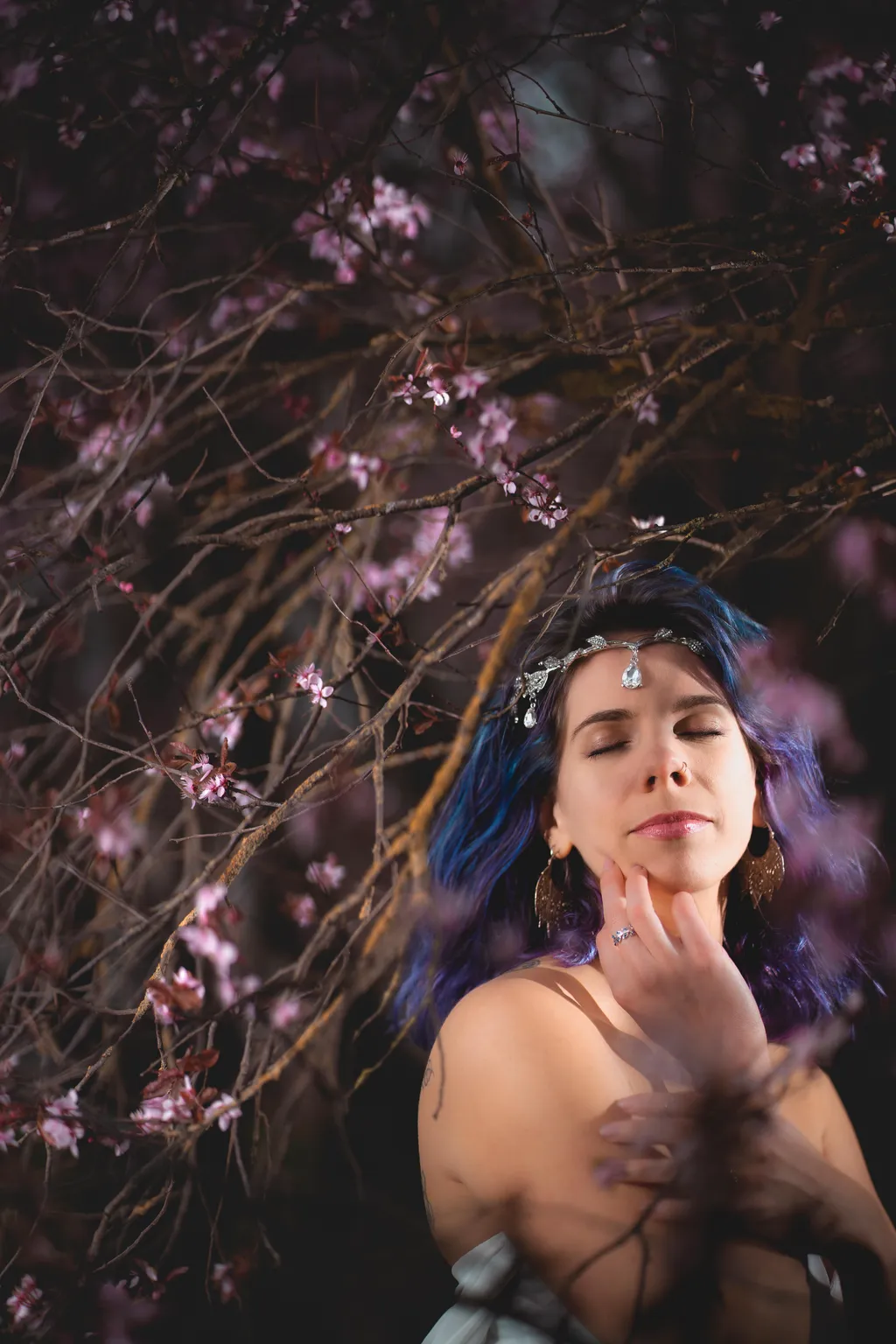 Editorial portrait in Luxembourg: woman with blue-purple hair resting hand on chin among soft pink spring blossoms