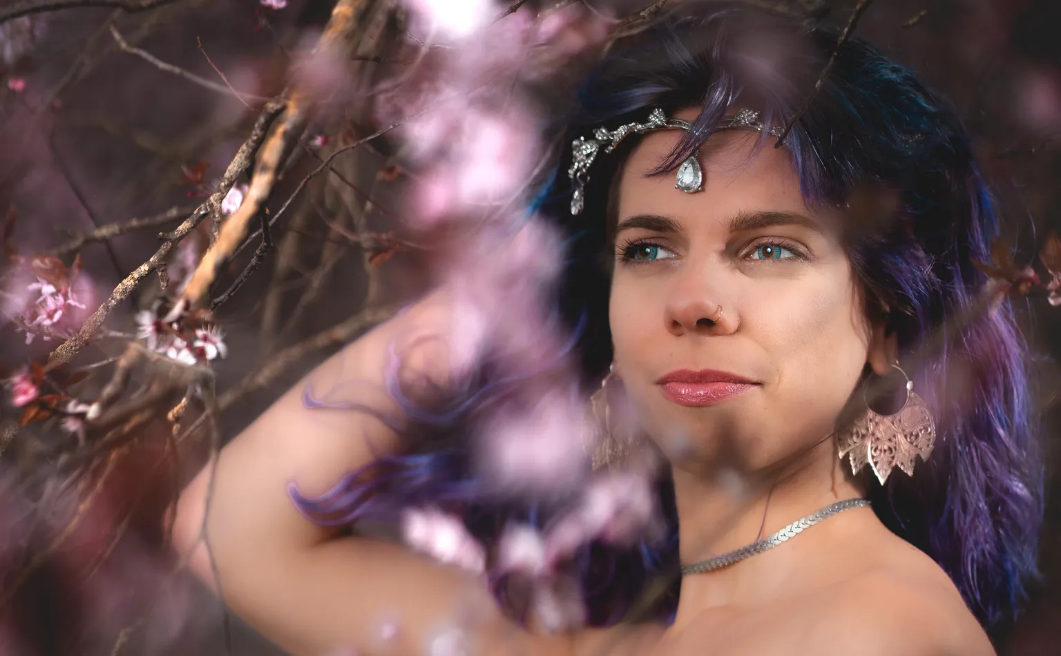 Close-up creative portrait of blue-eyed model in a teardrop headpiece, blossoms blurred across the foreground