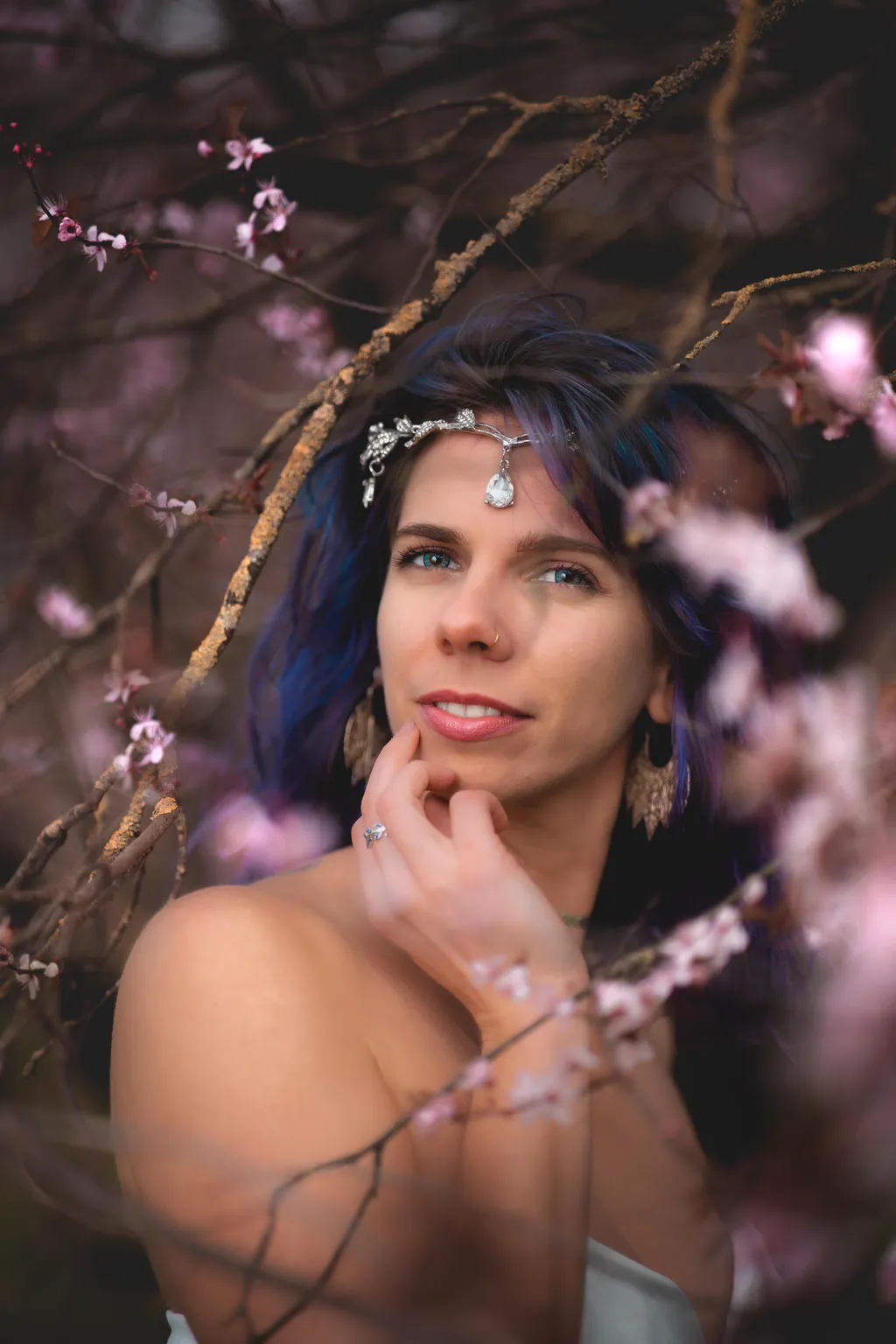 Editorial portrait of a model with blue-purple hair and silver jewellery gazing past pink plum blossoms