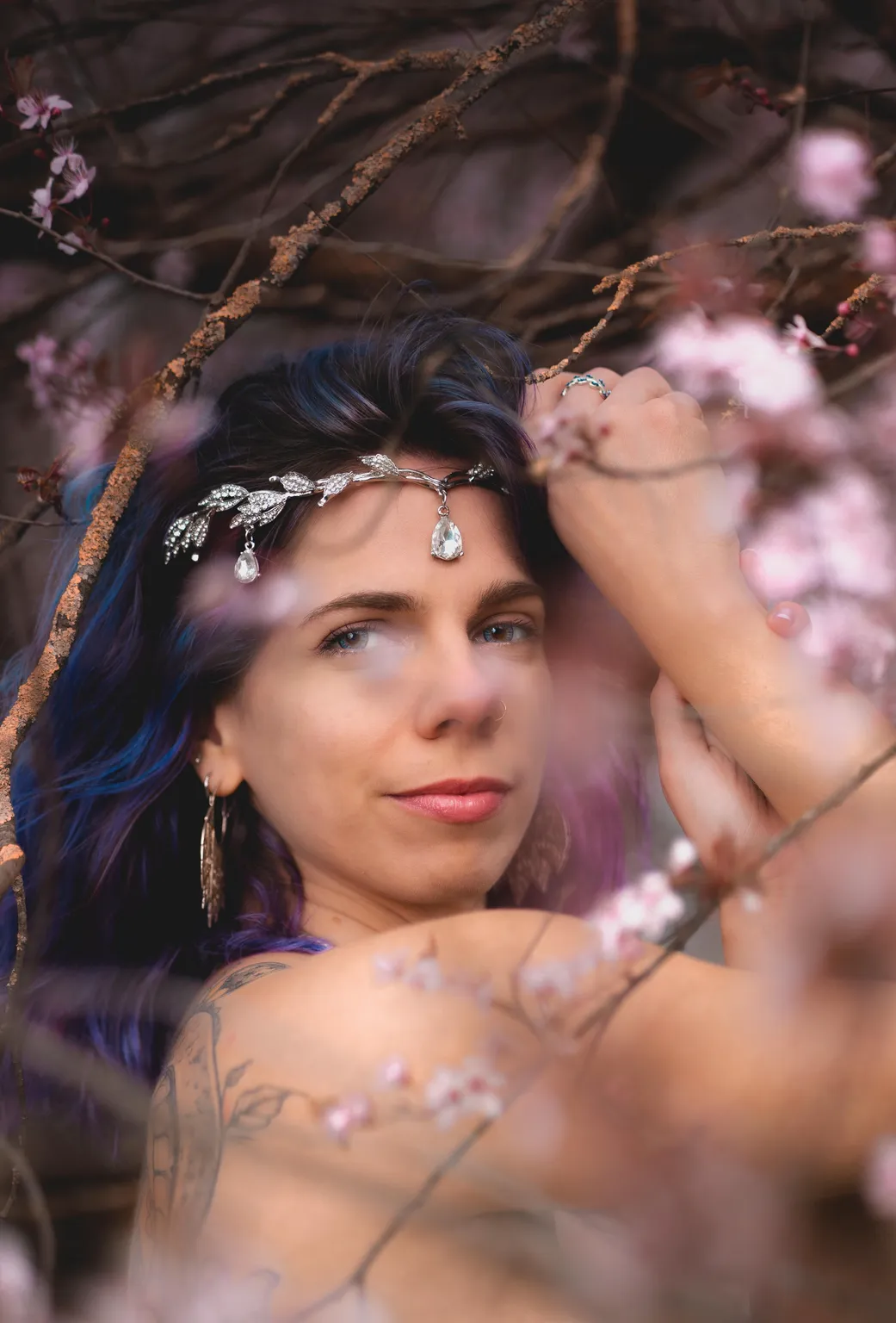 Intimate creative portrait of a model reaching into a blossoming branch, shoulder tattoo and silver crown visible