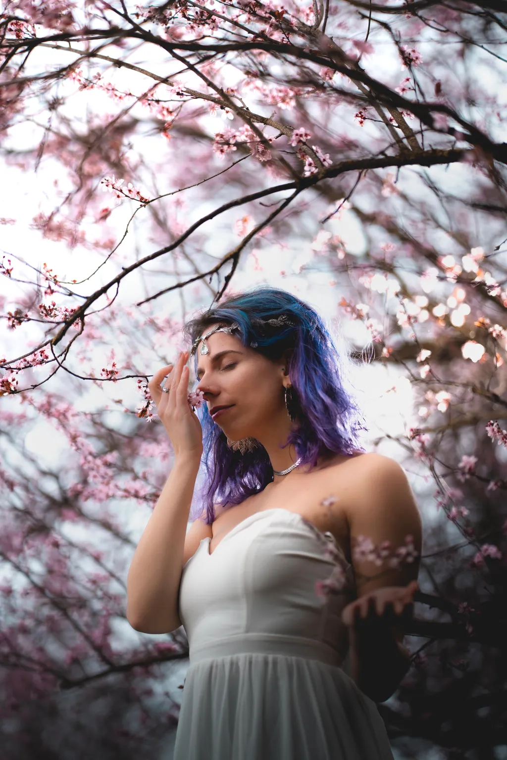 Backlit Luxembourg portrait: model in white gown breathing in pink blossoms against a bright spring sky