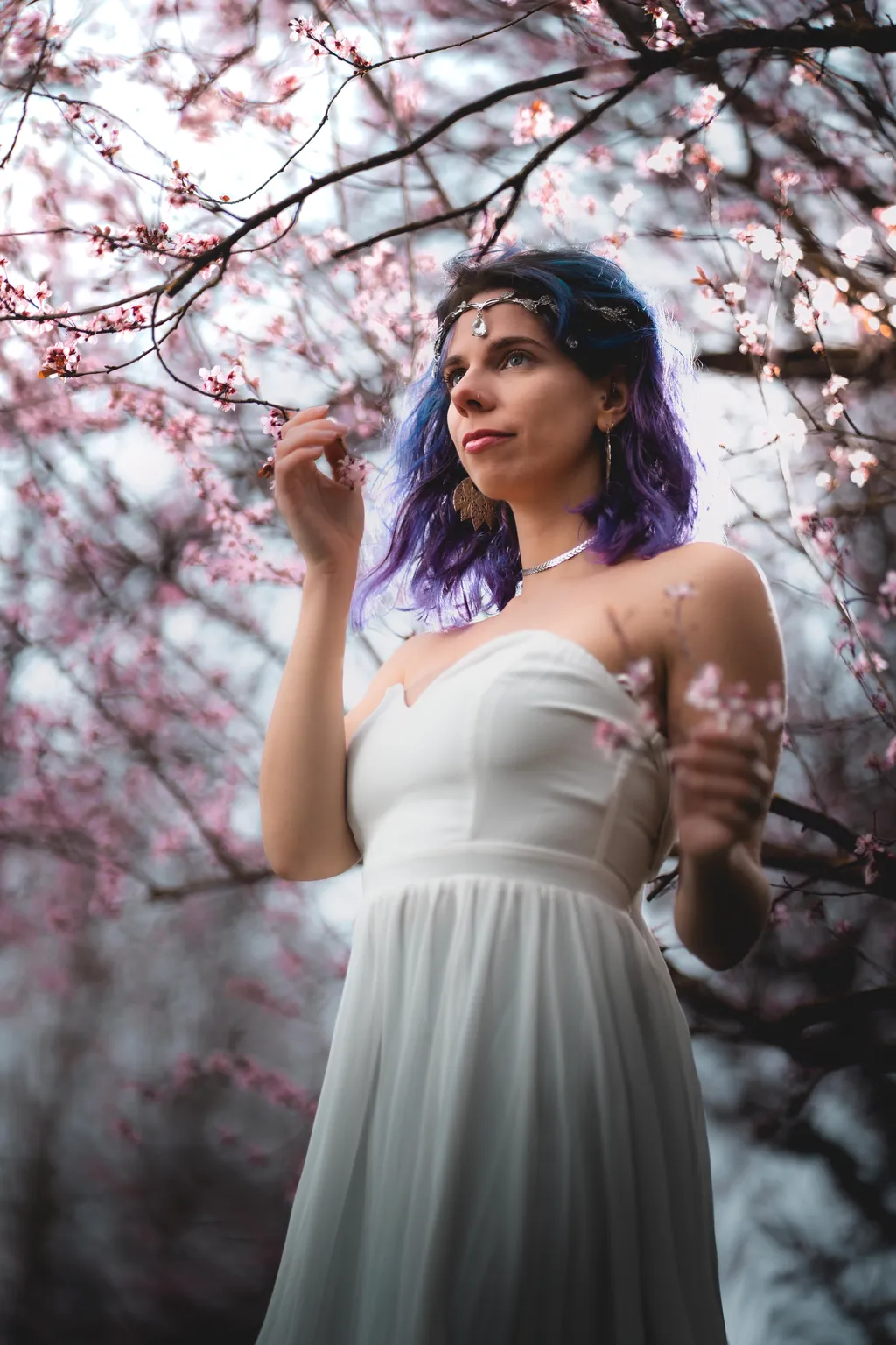 Editorial spring portrait of a woman in a flowing white dress holding a delicate blossom-laden branch