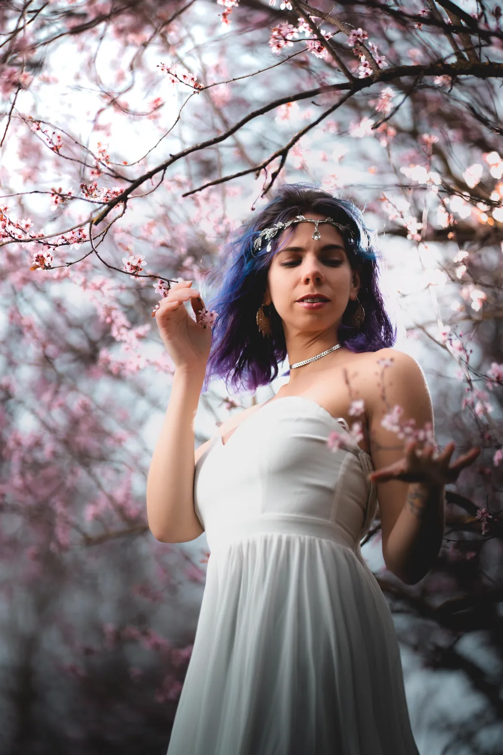 Creative portrait of a blue-haired model in a white gown, hands raised among cascading pink blossoms