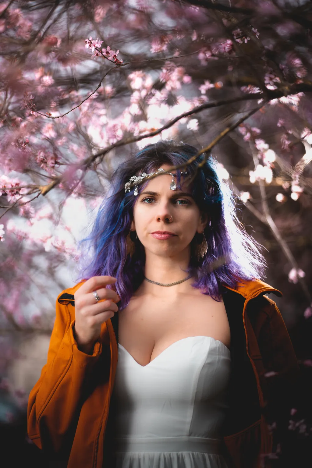 Striking portrait of a woman in an orange jacket over a white gown, framed by dense pink blossom branches