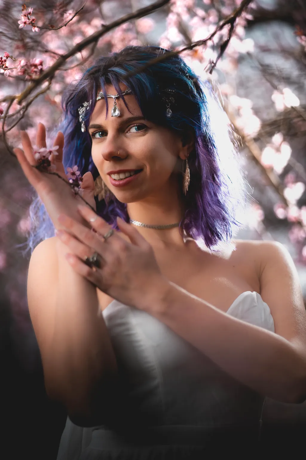 Warm editorial portrait of a smiling woman in a white gown, hands clasped beside a blossoming branch
