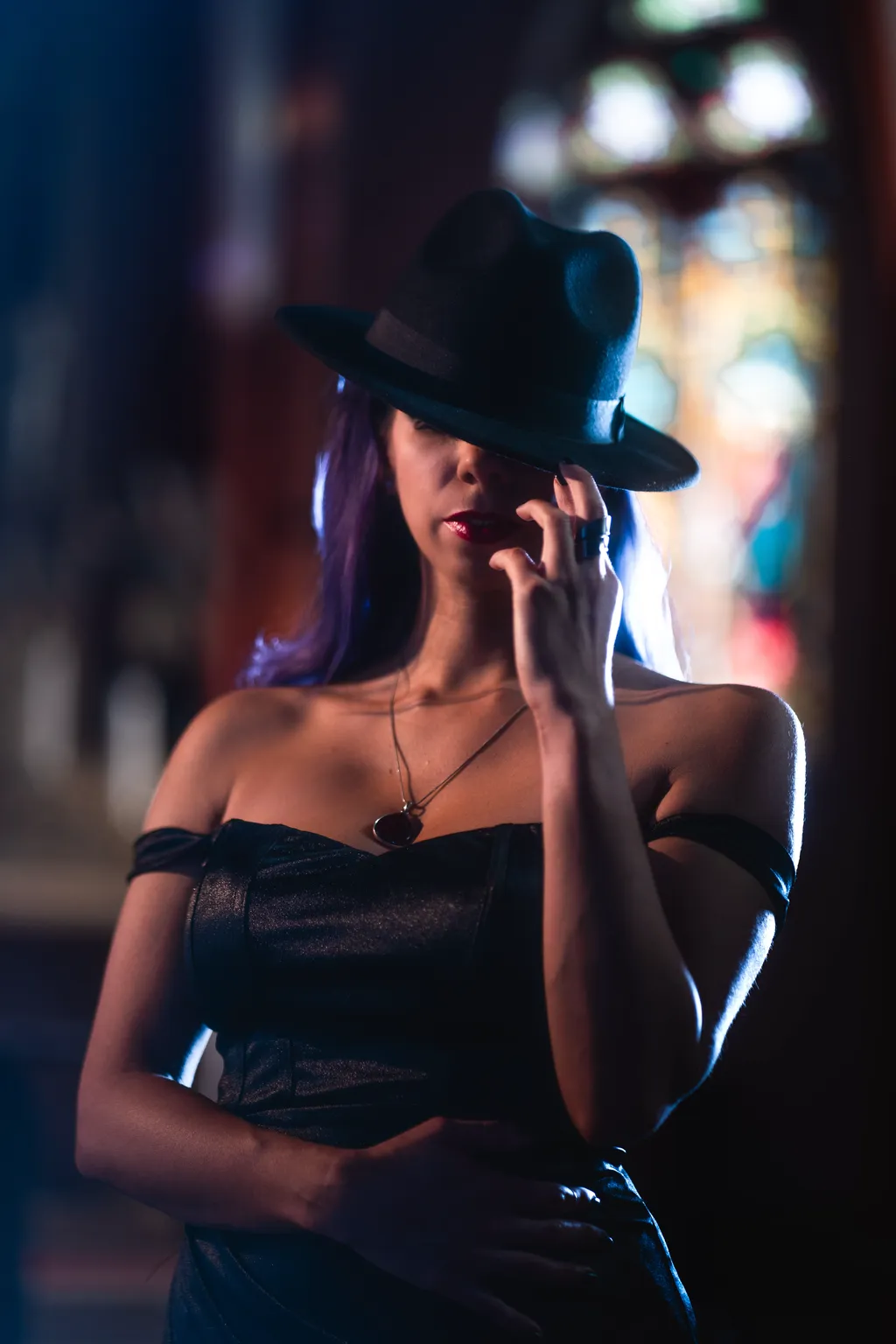 Close-up fine-art portrait of a woman in a black off-shoulder gown and hat against stained glass, shot in Luxembourg